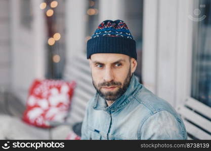 Good looking European male with thick beard and mustache wears hat and denim jacket, has thoughtful expression, contemplates about something, poses indoor. Pensive young bearded stylish man.