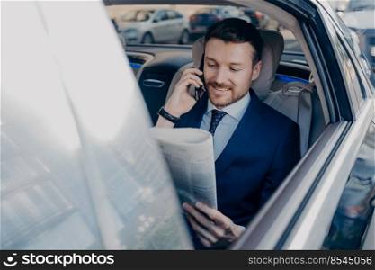 Good looking happy executive manager in stylish formal wear reads newspaper on backseat of car in same time speaking on smartphone with business partner, while staying in road traffic. Good looking young executive manager in formal wear reads newspaper on backseat of car