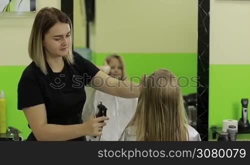 Good looking little girl getting her hair wet before getting a haircut in beauty salon. Beautiful female barber spraying water on client&acute;s hair in hairdressing salon.