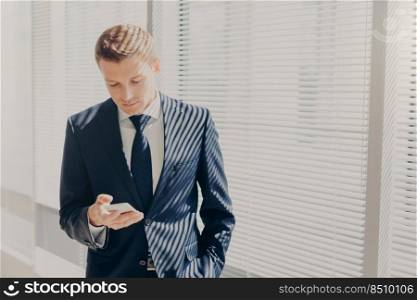 Good looking male executive dressed in corporate clothing, types text message, stands in office building, types feedback on mobile phone, stands near window at his cabinet. Technology concept