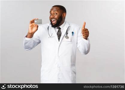 Good-looking pleased male doctor, physician in white coat, holding credit card and show thumb-up in approval or like, recommend bank, online purchases or contactless payment for hygiene purpose.. Good-looking pleased male doctor, physician in white coat, holding credit card and show thumb-up in approval or like, recommend bank, online purchases or contactless payment for hygiene purpose