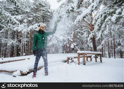 Good looking positive male keeps hand on fir tree, looks with cheerful expression, enjoys beauty of trees covered with snow, spends weekends in forest. Smiling bearded man being in good mood
