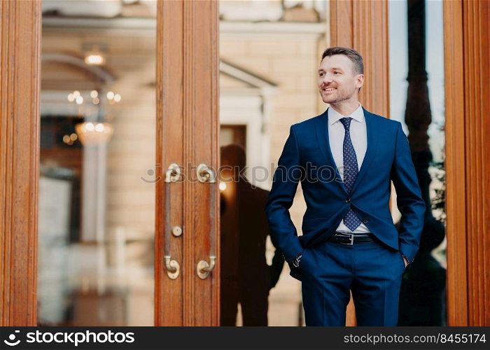 Good looking satisfied business worker wears elegant luxury suit, stands near entrance to restaurant, waits for colleagues, celebrate something, looks happily aside, has positive expression.