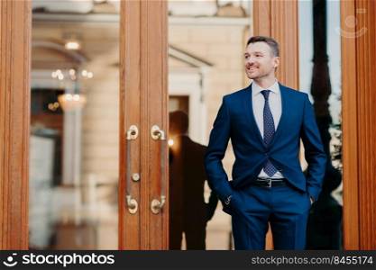 Good looking satisfied business worker wears elegant luxury suit, stands near entrance to restaurant, waits for colleagues, celebrate something, looks happily aside, has positive expression.
