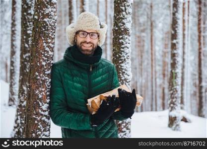 Good looking smiling male wears warm winter clothes holds firewood, stands near tree, spends free time with friends in winter forest, going to have picnic, makes fire to warm in frosty weather