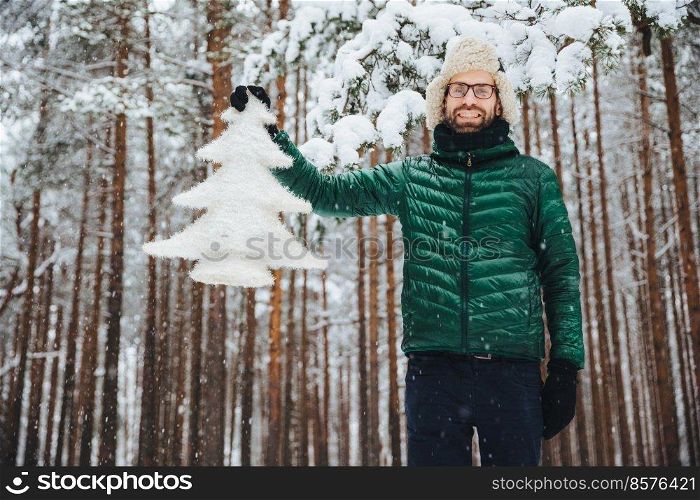 Good looking unshaven male in eyewear, wears warm winter clothes, holds white fir tree, has good mood, being in anticipation of New Year and Christmas, poses against winter high trees