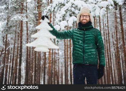 Good looking unshaven male in eyewear, wears warm winter clothes, holds white fir tree, has good mood, being in anticipation of New Year and Christmas, poses against winter high trees