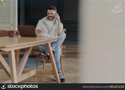 Good looking young businessman in casual wear talking about work or business on mobile phone and looking at monitor on laptop while sitting at wooden desk in modern luxury office or apartment. Young laughing man freelancer talking on mobile phone while sitting at his workplace at home, working remotely on laptop