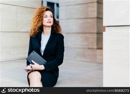 Good-looking young businesswoman with luxuriant hair, blue eyes, red lips, wearing black suit, holding in hands pocket book and modern tablet computer, looking aside with thoughtful expression