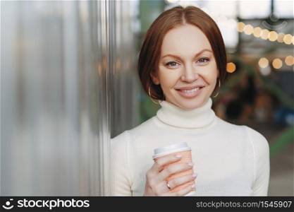 Good looking young female dressed in casual white turtleneck sweater, holds paper cup of aromatic cappucino or coffee, looks happily at camera, poses outside, has spare time. Lifestyle concept