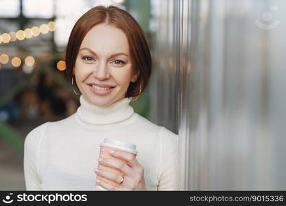 Good looking young female dressed in casual white turtleneck sweater, holds paper cup of aromatic cappucino or coffee, looks happily at camera, poses outside, has spare time. Lifestyle concept