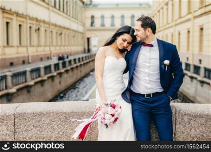 Good looking young female with appealing appearance wears white wedding dress, holds nice bouquet, leans at lover`s shoulder, enjoy togetherness and calm atmosphere as stand on bridge alone.