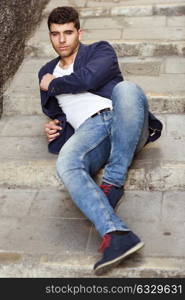 Good looking young man with blue eyes in the street. Model of fashion sitting in urban stairs wearing white t-shirt, jeans and blue jacket