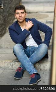 Good looking young man with blue eyes in the street sitting on steps. Model of fashion in urban background wearing white t-shirt, jeans and blue jacket