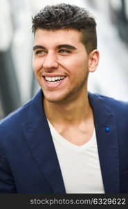 Good looking young man with blue eyes smiling in the street. Model of fashion in urban background wearing white t-shir and blue jacket