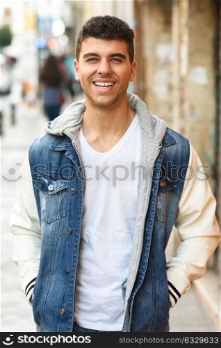 Good looking young man with blue eyes smiling in the street. Model of fashion in urban background wearing white t-shirt, jeans and blue jacket
