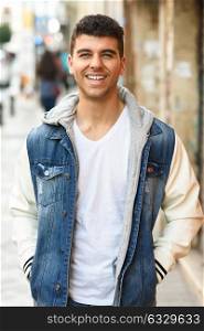 Good looking young man with blue eyes smiling in the street. Model of fashion in urban background wearing white t-shirt, jeans and blue jacket