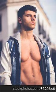 Good looking young man with muscular body in the street. Model of fashion in urban background wearing jeans and blue jacket