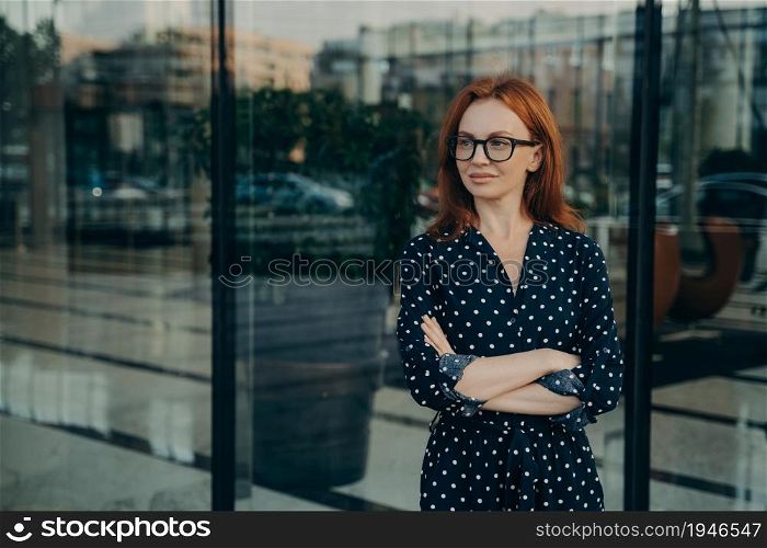 Good looking young redhead woman keeps arms crossed looks thoughtfully away being deep in thoughts wears transparent glasses polka dor dress stands near office building window waits for someone. Good looking young redhead woman stands near office building window
