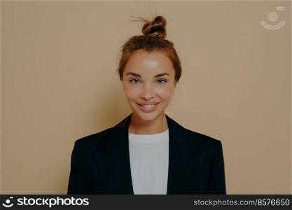 Good looking young woman with delighted face expression wearing elegant formal suit and cheerfully smiling at camera, ready for business meeting on beige background. Human face expression. Young woman ready for business meeting on beige background