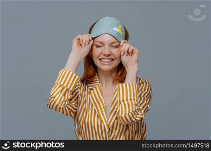 Good morning, lifestyle. Smiling freckled woman takes off blindfold, wears yellow striped pajama, awakes with smile after seeing pleasant dreams, feels relaxed and refreshed, poses over grey wall