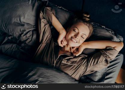 Good morning. View from above of young beautiful relaxed brunette female with closed eyes in satin pajamas waking up in her bed fully rested, happy woman stretching after night sleep. Young beautiful relaxed brunette female in satin pajama stretching in bed
