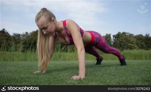 Gorgeous blonde woman with pony tail warming up and doing push ups. Girl working out on green grass crossfit strength training on nice summer day in the park