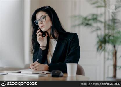Gorgeous businesswoman makes phone call, concentrated in computer screen, writes down information into notepad, poses at cozy workplace, focused on important task, makes notes in personal organizer