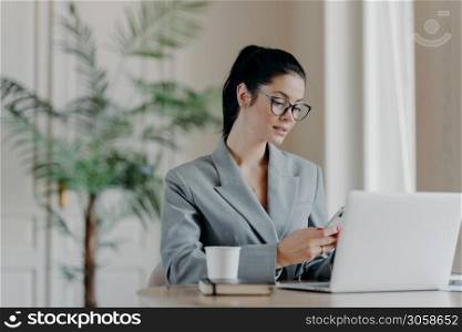 Gorgeous busineswoman types message on smartphone, works on new publication, poses in front of laptop computer, involved in working process, sits at desktop, drinks aromatic coffee, uses internet