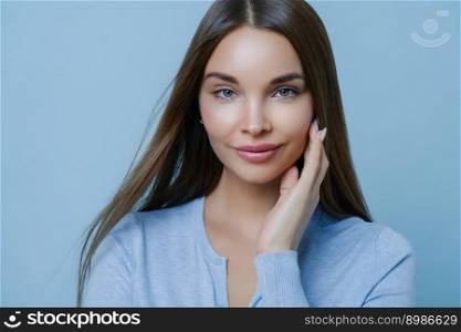 Gorgeous dark haired woman touches cheek gently, has direct gaze at camera, looks with tenderness, wears casual sweater, poses against blue background, has perfect glowing skin. Beauty, hair care