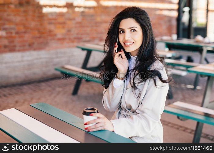 Gorgeous dark-haired woman with pure skin, dark bright eyes and full lips holding takeaway coffee and smartphone chatting with her boyfriend having pleased look. Young female enterpreneur resting