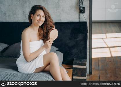 Gorgeous european woman combs her long silky hair with hairbrush and smiling. Happy girl wrapped in towel doing hair treatment after bathing. Young woman takes morning shower at home and doing hair.. Gorgeous european woman wrapped in towel combs her long silky hair with hairbrush and smiling.