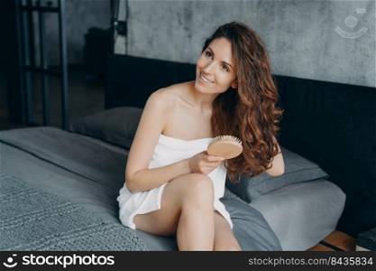 Gorgeous european woman combs her soft curly hair with hairbrush. Girl wrapped in towel doing hair treatment after bathing. Young woman takes shower at home and doing hair. Weekend morning at home.. Weekend morning at home. Gorgeous european woman combs her soft curly hair with hairbrush.