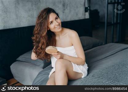 Gorgeous european woman combs her soft curly hair with hairbrush. Girl wrapped in towel doing hair treatment after bathing. Young woman takes shower at home and doing hair. Weekend morning at home.. Weekend morning at home. Gorgeous european woman combs her soft curly hair with hairbrush.
