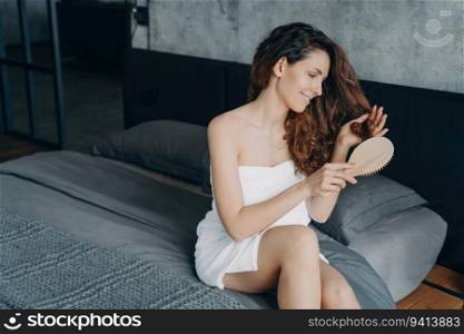 Gorgeous European woman combs long silky hair in bedroom. Hair cosmetics and treatments post-bath. Young woman showers, styles hair in morning. Femininity and beauty concept.