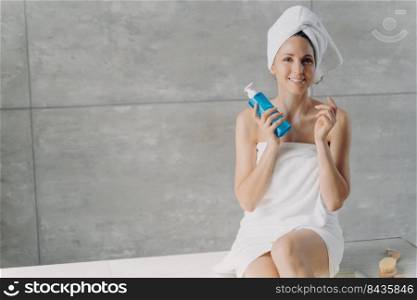 Gorgeous european woman wrapped in towel after bathing. Face toner advertising concept banner on copy space. Young woman is smiling holding bottle of micellar water. Face washing foam in bottle.. Gorgeous european woman holding bottle of micellar water. Face washing foam in bottle.