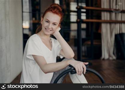 Gorgeous ginger female fitness instructor in white t-shirt smiles at camera during Pilates ring workout in gym. Promoting a healthy, active lifestyle.