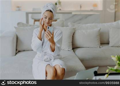 Gorgeous lady touches face gently applies face cream makes video call holds smartphone in front of herself wears bathrobe and towel sits on comfortable sofa against cozy home interior. Beauty concept