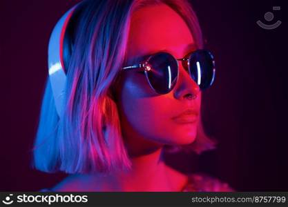Gorgeous lady with dyed hair and sunglasses listening music in headphones and singing with neon light background. Charming hipster girl dancing with eyes closed. Gorgeous lady with dyed hair and sunglasses listening music in headphones and singing with neon light background. Charming hipster girl dancing with eyes closed.