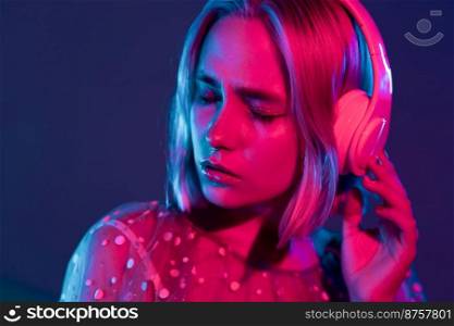 Gorgeous lady with dyed hair listening music in headphones and singing with neon light background. Charming hipster girl dancing with eyes closed. Gorgeous lady with dyed hair listening music in headphones and singing with neon light background. Charming hipster girl dancing with eyes closed.