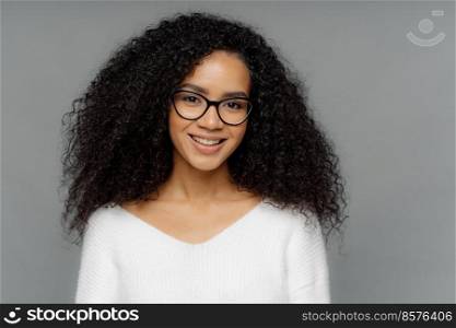 Gorgeous lovely curly woman with Afro hairstyle, feels glad, smiles gently at camera, wears optical glasses and white sweater, isolated on grey background. Happy emotions and feelings concept