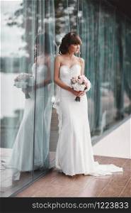 gorgeous luxury stylish bride in a white dress with a bouquet in hands near the panoramic window on the cloudy sky background. gorgeous luxury stylish bride in a white dress with a bouquet in hands near the panoramic window