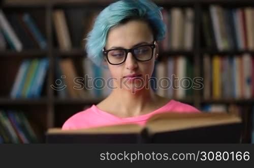 Gorgeous nerdy college female student in trendy eyeglasses reading a book in university library. Young hipster woman with blue hair researching in library. Serious student preparing for exam.