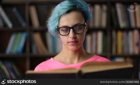 Gorgeous nerdy college female student in trendy eyeglasses reading a book in university library. Young hipster woman with blue hair researching in library. Serious student preparing for exam.