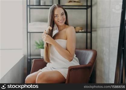 Gorgeous pretty young woman has well cared long healthy shiny hair, combes with hair brush, sits in comfortable armchair wrapped in bath towel, poses against bathroom interior. Beauty concept