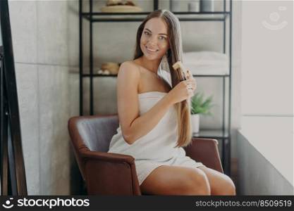 Gorgeous pretty young woman has well cared long healthy shiny hair, combes with hair brush, sits in comfortable armchair wrapped in bath towel, poses against bathroom interior. Beauty concept