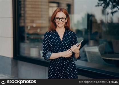 Gorgeous redhead businesswoman walks in urban place holds tablet and diary strolls near restaurant during daylight hours wears polka dot dress spectacles looks happily into distance walks in city. Redhead businesswoman walks in urban place holds tablet and diary