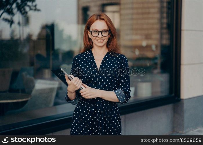 Gorgeous redhead businesswoman walks in urban place holds tablet and diary strolls near restaurant during daylight hours wears polka dot dress spectacles looks happily into distance walks in city. Redhead businesswoman walks in urban place holds tablet and diary