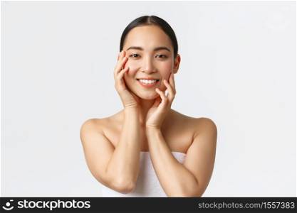 Gorgeous sensual asian woman in towel touching face and smiling, applying skincare products, cosmetic procedure at spa salon, massaging face and gazing camera happy, white background.. Gorgeous sensual asian woman in towel touching face and smiling, applying skincare products, cosmetic procedure at spa salon, massaging face and gazing camera happy, white background