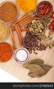 gorgeous setting with cooking spices and herbs (bay leaves, chili powder, coriander, cloves, cardamom pods, cinnamon sticks, garam masala, piri piri, salt, turmeric) on a wooden mat